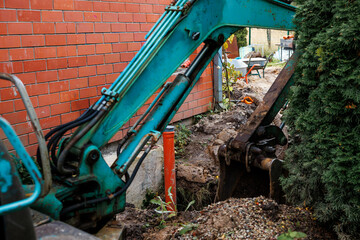 A blue excavator digs near a red brick wall with an orange pipe exposed. Gravel, tools, and a tree partially obscuring the bucket are visible.