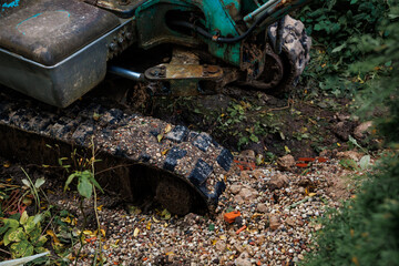 A small excavator's tracks and lower body, covered in dirt and mud, rest on gravel and soil with scattered vegetation and orange debris fragments.