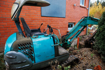 A blue excavator with an open canopy rests near a red brick building, its arm extended into a soil filled area. A bush partially obscures the frame.