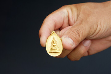 Close up hand hold gold Buddhist amulet with a sitting Buddha image on a dark background,...