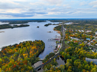 High angle panoramic view of a railway bridge crossing water, a marina, and a town nestled amongst a vibrant autumn forest and wide river
