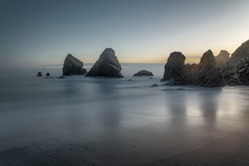 Long-exposure Irish beach with dark sand, towering sea stacks, and smooth misty water at twilight.