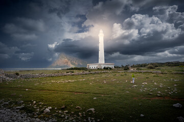 High white lighthouse on coast with sunlit mountain against dramatic dark cloudy sky, Sicily, Italy