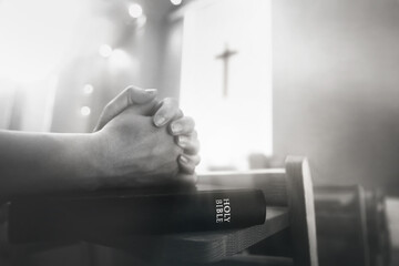A believer praying with both hands together during a church service, with the Holy Bible and the cross of Jesus Christ in the background.
