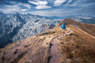 Fototapeta premium Female hiker walks narrow path to sunlit hilltop mountain ridge sunny day, Prokletije, Montenegro