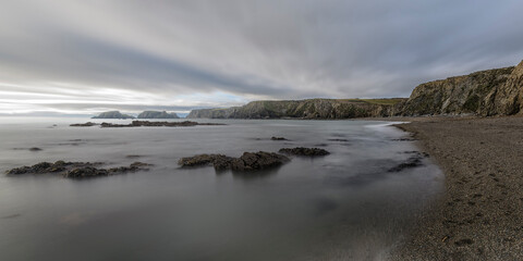 Panoramic Irish cove with smooth long-exposure water, dark pebbled beach, and dramatic cliffs under streaked clouds.