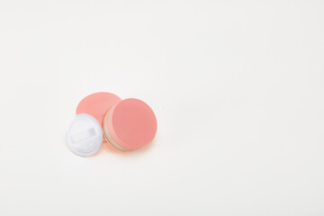Three pink and white makeup items are on a white background. The makeup items are small and circular, and they appear to be powder or cream. Scene is light and playful.