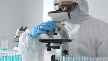 Medical virologist in protective hazmat gear examining viral samples under a microscope while...