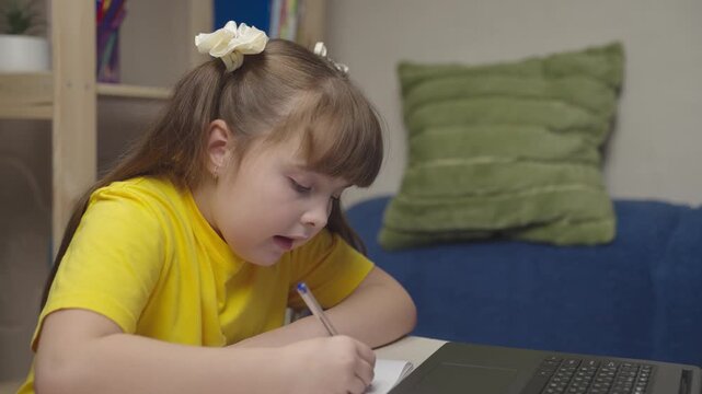 little girl child does homework while sitting table. kid writes with pen notebook. modern laptop smart child learning. modern education kid home. daughter does homework children room. happy family