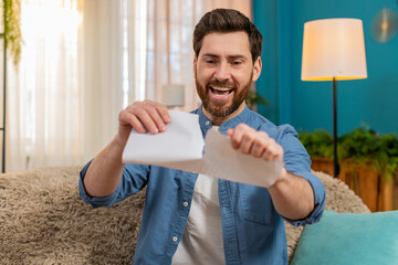 Young man at home sits on the sofa, smiling and tearing an unnecessary document into pieces with...