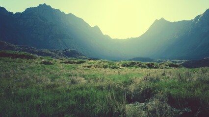 Golden sunlight bathes the vast landscape, illuminating the towering mountains and vibrant green fields. Nature awakens to a beautiful dawn, creating a serene and magical atmosphere.