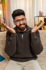 Indian man at home sits at sofa and spreads hands with confused face, reacting to unclear message....