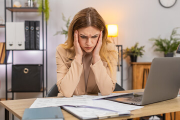 Middle-aged businesswoman at home office looks at bills invoices receipt on laptop sees costs go up frowns confused and unhappy. Freelancer girl at table writes savings plan sets targets prints page