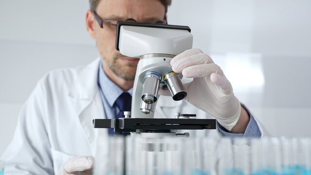 Scientific researcher wearing lab coat, glasses and white protective gloves carefully calibrating laboratory microscope, performing precision medical investigation. Medicine, healthcare and science - Powered by Adobe