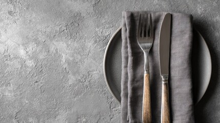 A gray ceramic plate sits next to a folded gray linen napkin, with a fork and knife laid atop. The background is a textured gray surface