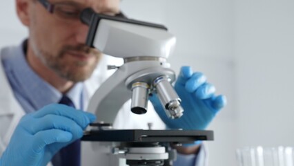 Close up of male scientist in white coat and blue gloves adjusting microscope, conducting pharmaceutical research in modern laboratory. Medicine and science concept