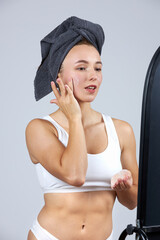 A young beautiful girl in white lingerie with a towel on her head stands in front of the mirror and does cosmetic procedures.