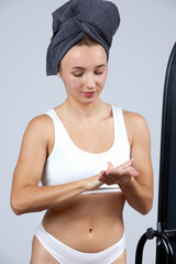 A young beautiful girl in white lingerie with a towel on her head stands in front of the mirror and does cosmetic procedures.