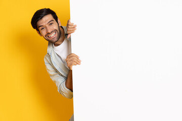 Happy young man is standing behind the white blank banner or empty copy space advertisement board isolated on yellow background