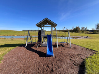 Children's play structure with blue slide and swings on mulch, surrounded by grassy fields, sparse trees, a pond with reeds, and a clear blue sky.