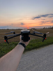 A person holds a drone with red lights illuminated, set against a sunset. An open field with harvested crops, a gravel path, and power lines are visible.