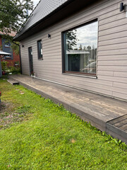 Modern house with light brown wooden siding, sloped roof, wooden deck, large windows, grassy area, and a potted plant near the entrance.