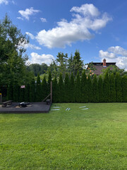 A well maintained lawn bordered by evergreen trees, a wooden deck with a bench and grill, and a house with a chimney visible in the distance.