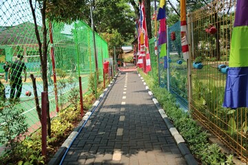 A sunlit walkway runs between bright fences, with colorful flags along one side and a sports court nearby. People enjoy the outdoor space, creating a lively, festive atmosphere.