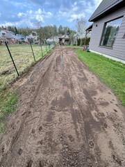 A muddy dirt path with visible tire tracks runs between a fenced area and a modern gray house. Additional houses and trees are visible in the distance.