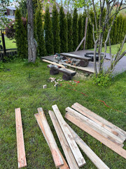 Residential backyard featuring a wooden deck, fire pit with surrounding logs, scattered planks on grass, and a row of tall evergreen trees.