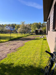 A wooden house with a bicycle leaning against it, a grassy yard, dirt path, fenced garden, and distant houses surrounded by trees under a clear sky.