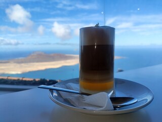 Traditional Canary Islands coffee Barraquito served at Mirador del Rio viewpoint on Lanzarote with scenic view of La Graciosa island and the Atlantic Ocean under clear blue sky.