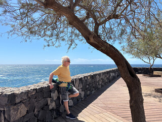 Senior white haired man standing along the sea relaxing and admiring the horizon - active pensioner enjoying nature and healthy lifestyle outdoors on a sunny windy day