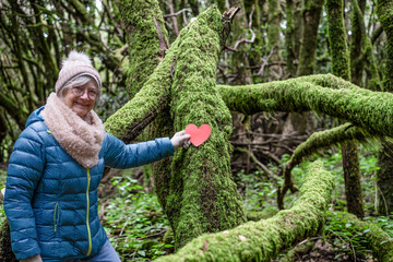 Smiling senior woman enjoying the freedom of the outdoors on a winter day walking in a mountain forest placing a red paper heart on the moss-covered branches.
