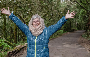 An active and smiling senior woman enjoys the freedom of the outdoors on a winter day, walking in a mountain forest with her arms outstretched