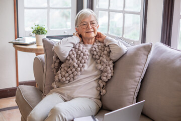 Smiling senior woman relaxing sitting comfortably on sofa in living room using computer, enjoying technology and social media