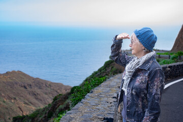 Smiling senior traveler woman in outdoor excursion looking at landscape and ocean view from top of hill enjoying vacation and travel. Horizon over water