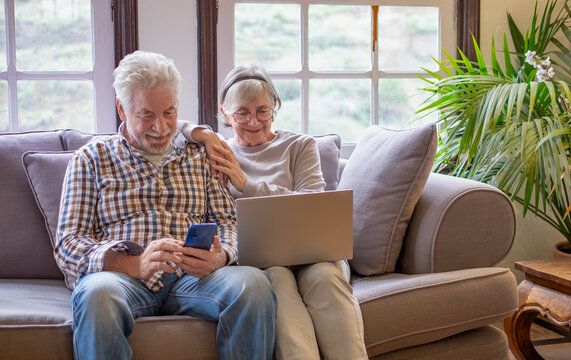 Smiling senior couple relaxing sitting on sofa together in living room using smartphone and computer, browsing internet on sofa at home enjoying tech and social
