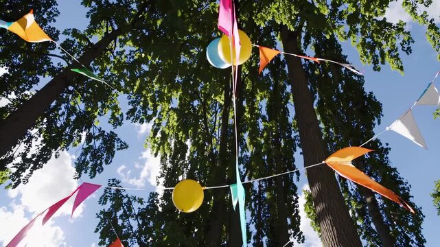 Colorful balloons and flags hang from tree. Outdoor pet celebration with party decor. Birthday park setup. Balloon and party banner. Pet party fun with balloons in sunny park, hat and fun vibe.