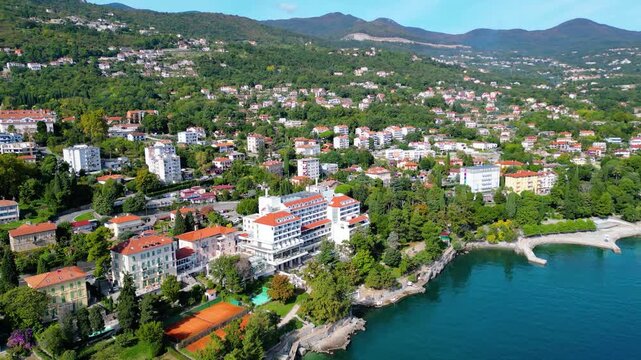 Aerial view of the Hotel Excelsior and the coastal promenade in Lovran, Croatia, surrounded by red-roofed houses, Adriatic Sea, and lush green hills of the Opatija Riviera