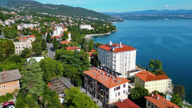 Aerial view of the Hotel Excelsior and the coastal promenade in Lovran, Croatia, surrounded by red-roofed houses, Adriatic Sea, and lush green hills of the Opatija Riviera