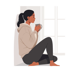Woman sits by the window, holding a mug, lost in thought, wearing a hoodie and leggings.