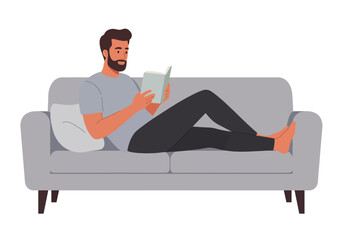 A man with a beard is relaxing on a gray couch, reading a book. He is barefoot and appears to be enjoying his leisure time.