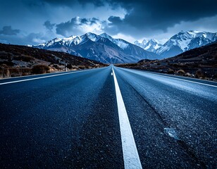 A long asphalt road stretches into the distance towards towering, snow-capped mountains under a dramatic, cloudy sky. The scene is cool toned