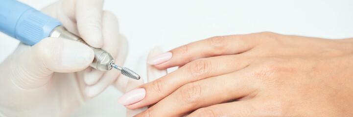 Close-up of manicure process on female hands with pink nails using electric nail file by gloved technician.