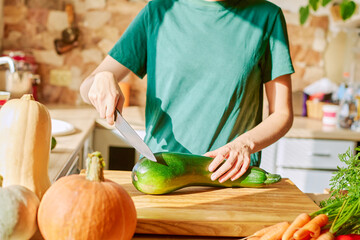 A woman in a green t-shirt is cutting a large green zucchini on a wooden board. Autumn background,...
