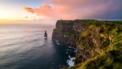 A majestic coastal cliff plunges into the sea, bathed in the soft light of a setting sun. The sky is painted with pastel hues