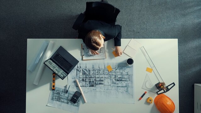 Aerial view of architect drawing while working at laptop. Top down view of caucasian engineer designing blueprint and drafting house model at table with equipment and safety helmet. Alimentation.