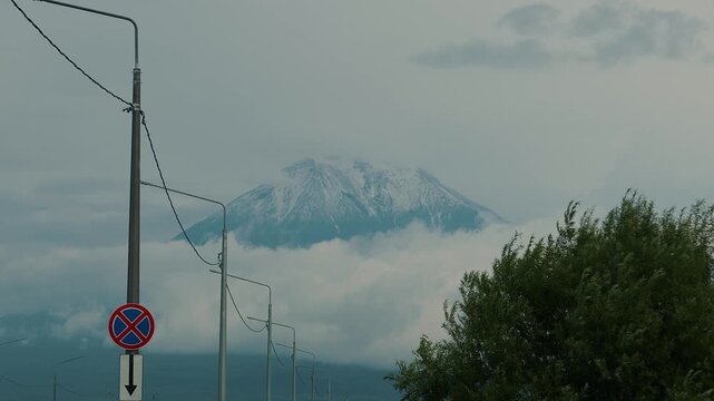 Breathtaking view of majestic mountain with snow-capped peak rising above clouds and row of lampposts with wires along road