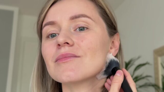 A 37-year-old woman applies bronzer with a makeup brush to contour her cheekbones, forehead, and neck. Close-up of fair skin, gray-blue eyes, light brown hair, natural daylight, and smooth blending.